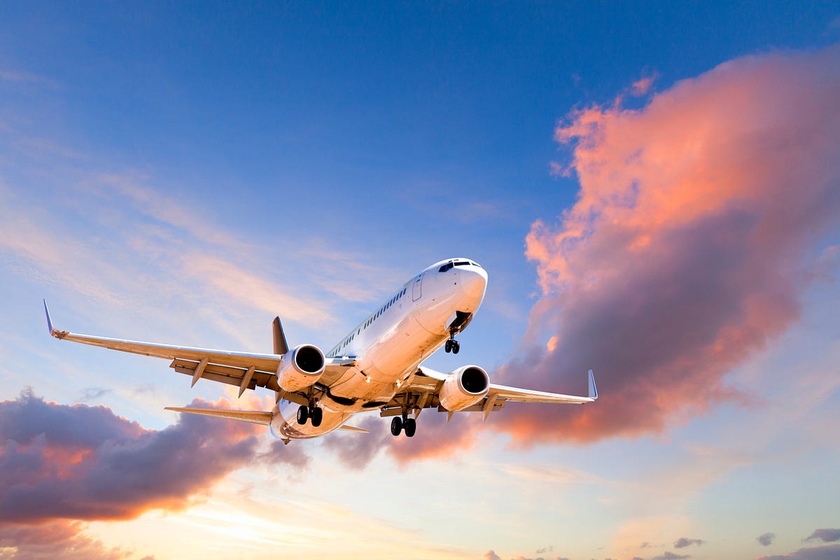 Commercial passenger jet taking off/ascending against a sunset sky with pink-orange clouds behind it, gear down in flight.